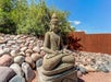 A meditating Thai Buddha statue in the center of a stone garden with trees and a fence behind it on a sunny day.