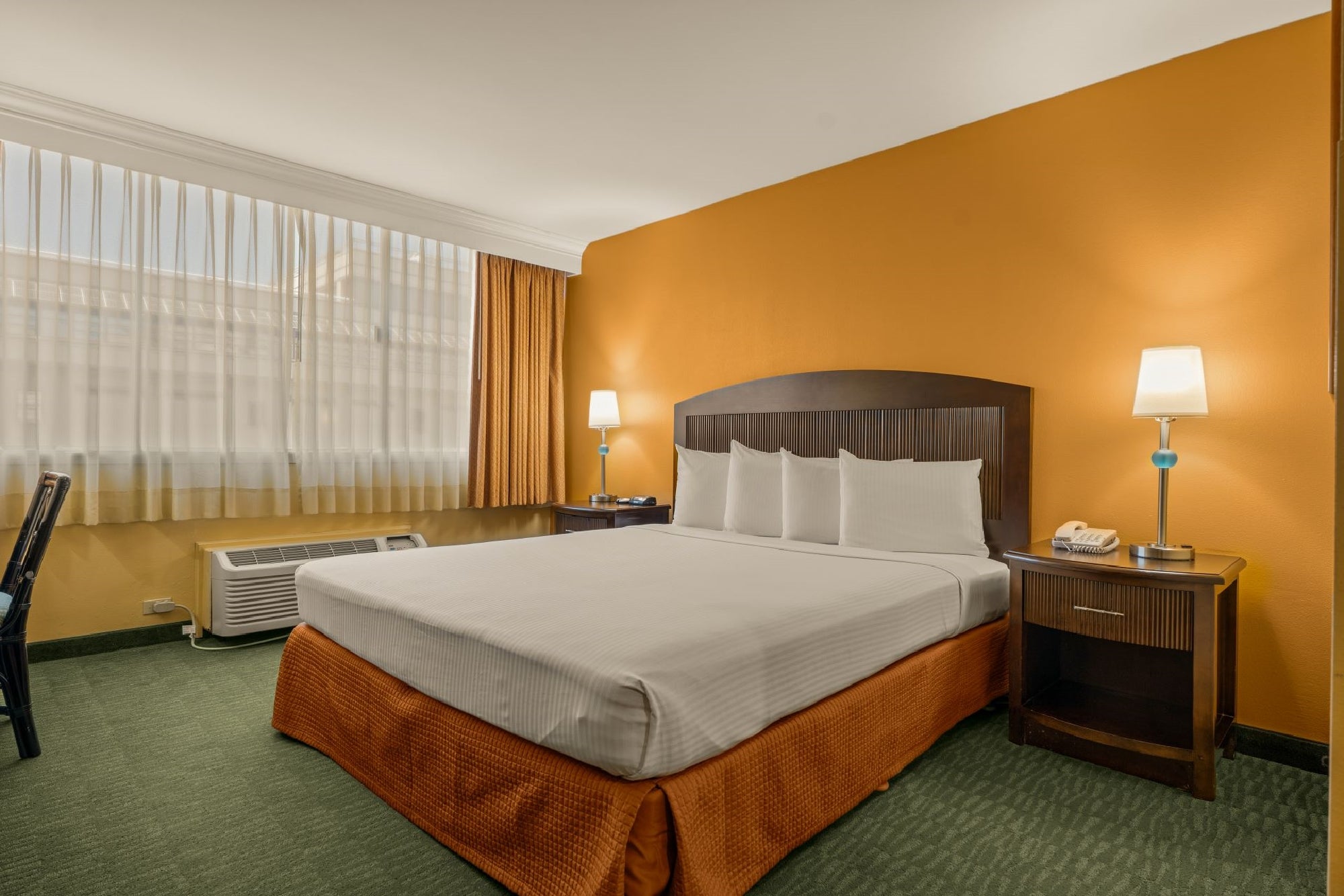 Hotel room with a king-sized bed, orange accent wall, green carpet, two bedside tables with lamps, a desk, and a window with curtains.
