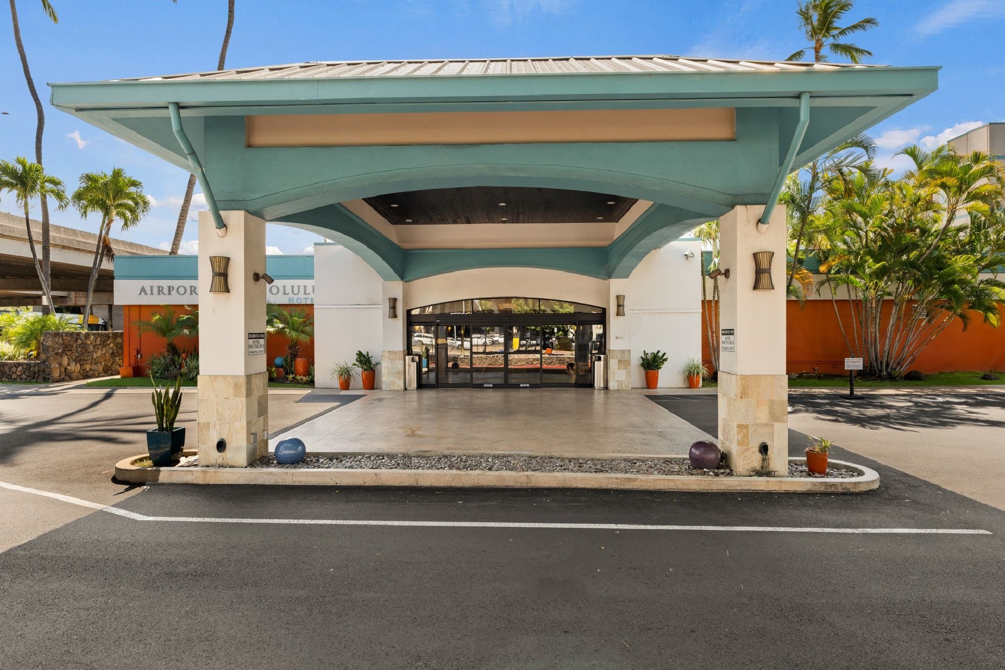 Covered entrance of a building with automatic glass doors, potted plants on both sides, and a driveway for vehicles.