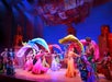 A colorful stage performance featuring dancers in vibrant costumes waving large, flowing fabrics under dramatic lighting.