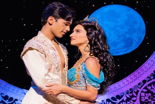 A man and woman in ornate costumes stand close together, gazing into each other's eyes, with a large blue moon and starry night sky in the background.