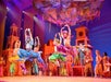 Performers in colorful costumes leap mid-air during a lively stage production with vibrant set pieces and dramatic lighting.