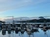 Several boats are docked at a marina with calm water, distant trees, and mountains under a partly cloudy sky in the background.