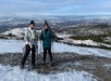 Two guests wearing cold-weather hiking attire and snowshoes and holding trekking poles as they smile at the camera with South Fork Campbell Creek valley in the background.