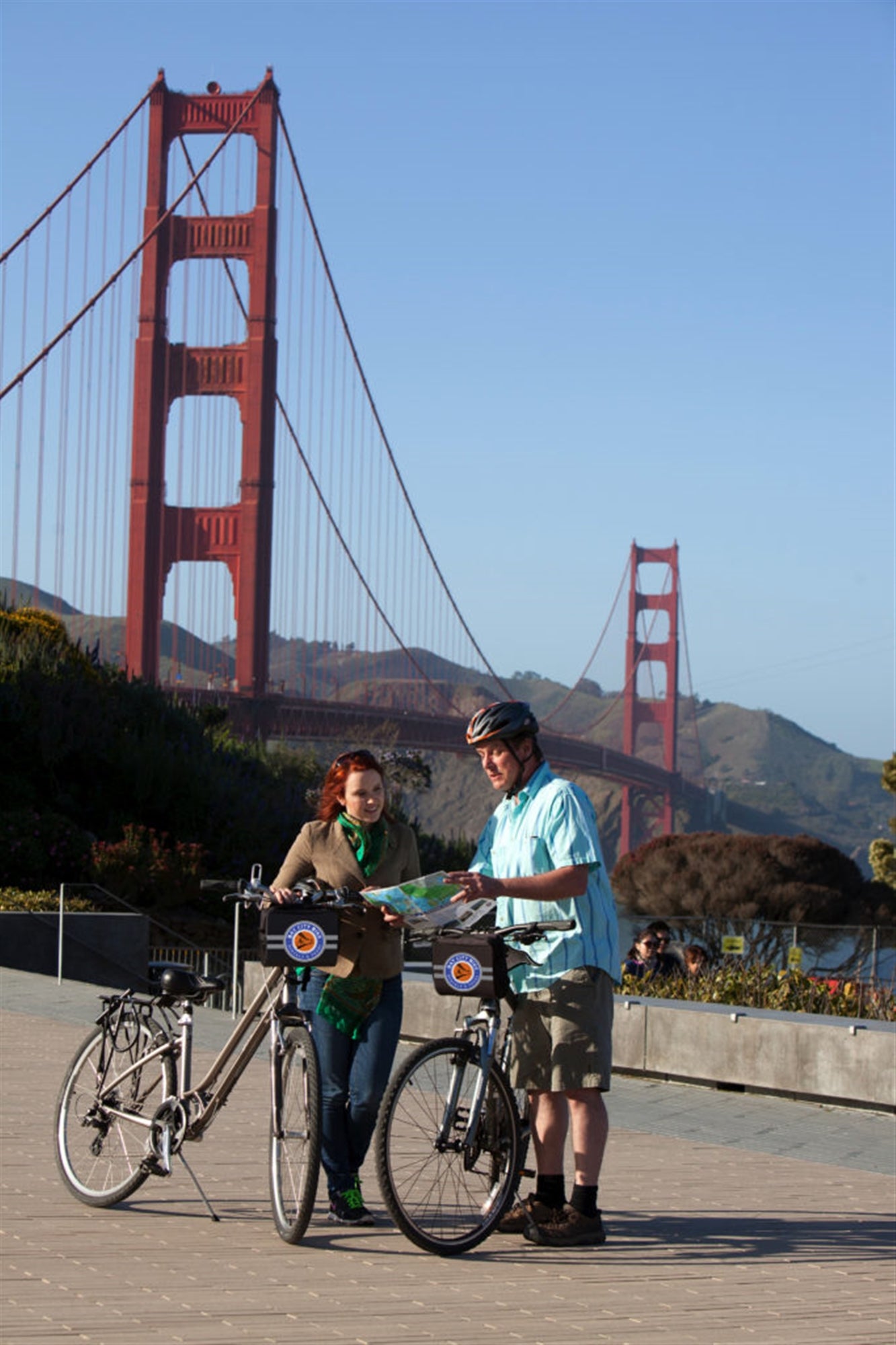 Two people with bicycles look at a map near the Golden Gate Bridge in San Francisco on a clear day.