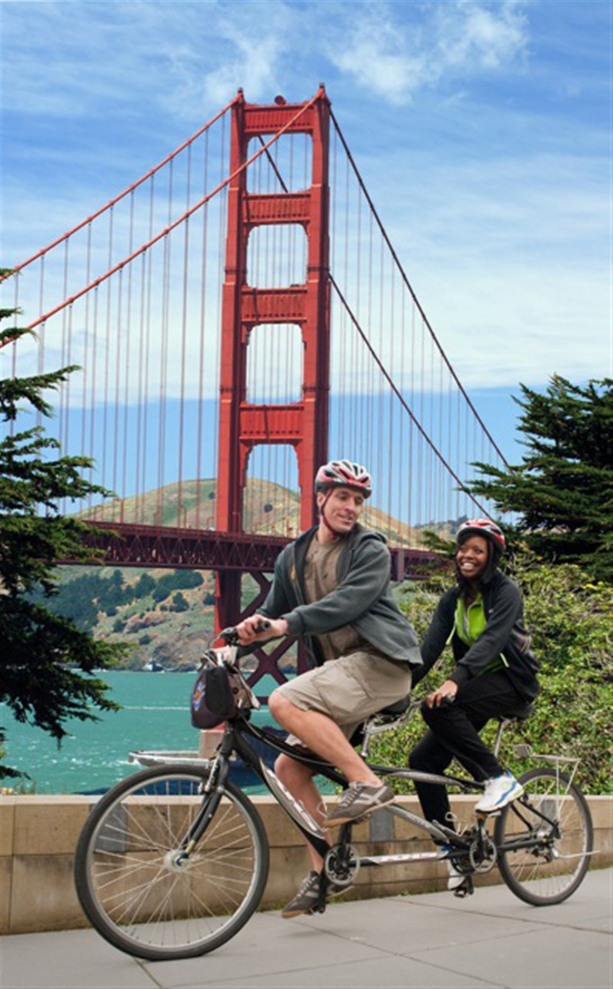 Two people ride a tandem bicycle in front of the Golden Gate Bridge on a clear day, both wearing helmets and casual clothing.