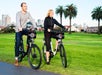 Two people ride electric bikes on a path in a park with city buildings and palm trees in the background.