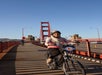 A woman wearing a helmet rides a bicycle on the Golden Gate Bridge bike path, with cars and another cyclist visible in the background.