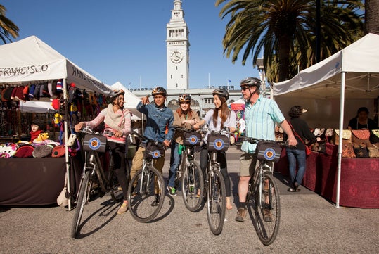 Group of five people with bicycles stand in front of a market area with tents, under a clear blue sky. A large clock tower is visible in the background.