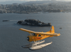 A yellow seaplane flies over water near Alcatraz Island, with a city skyline and mountains in the background.