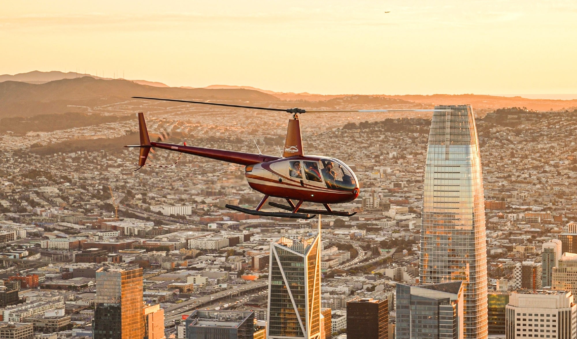 A red helicopter flies above a cityscape with tall buildings at sunset.