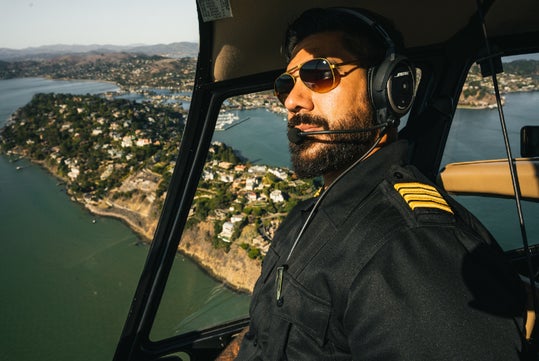 Helicopter pilot wearing sunglasses and a headset, flying over a coastal area with houses and hills in the background.