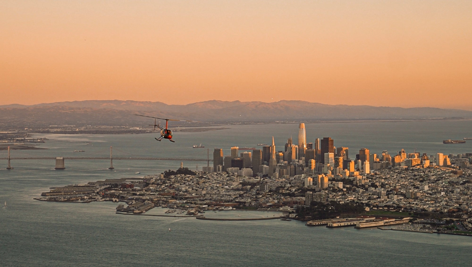 A gyrocopter flies over a city skyline with tall buildings at sunset, with a bay and distant mountains in the background.