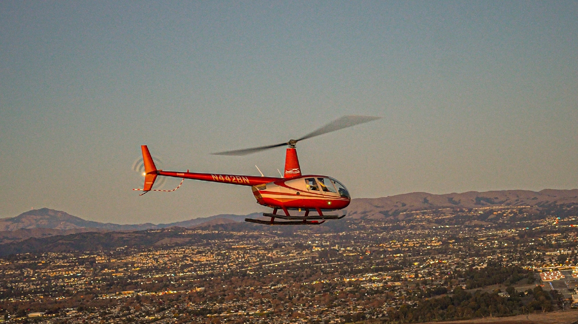 A red helicopter flies over a cityscape with mountains in the background under a clear sky.