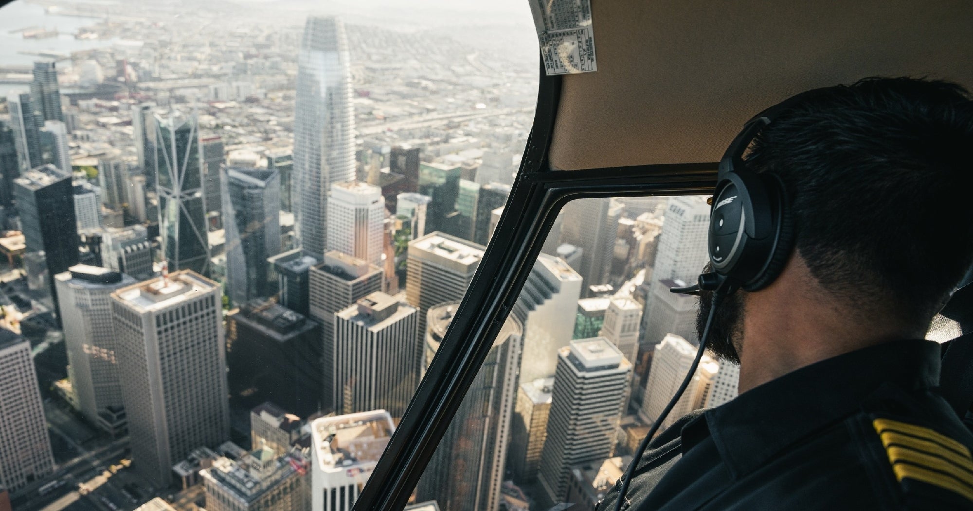 Pilot wearing a headset, flying a helicopter over a city with tall skyscrapers visible through the cockpit window.