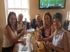 Five women sit around a wooden table at a bar, holding drinks and smiling. A TV above them displays a football game.