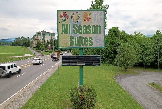 Sign for "All Season Suites" beside a road with vehicles and greenery, and a building in the background.