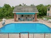 Outdoor swimming pool with turquoise water, surrounded by a red fence. A pool house with seating and decor is in the background, set amidst trees and greenery.