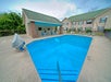 Outdoor swimming pool with railing, surrounded by a concrete deck. There are chairs and tables under a shaded area. Two-story building with green roof in the background.
