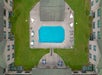 Aerial view of a rectangular outdoor swimming pool surrounded by grass and symmetric buildings. Umbrellas and lounge chairs are positioned around the pool area.