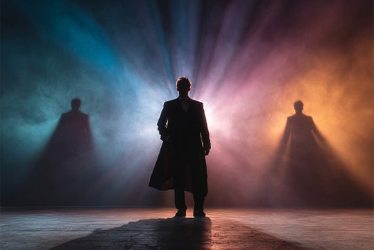 A person stands in the center of a stage with colorful lights and two shadows cast on a smoky backdrop, creating a dramatic atmosphere.