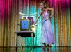 A woman in a formal dress performs a stage magic act, preparing to insert a sword into a blue box on a table in front of a gold curtain with colorful lighting.