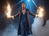 A man in a long coat stands on stage holding flames in both hands, surrounded by smoke and dramatic spotlights, with an audience in the background.
