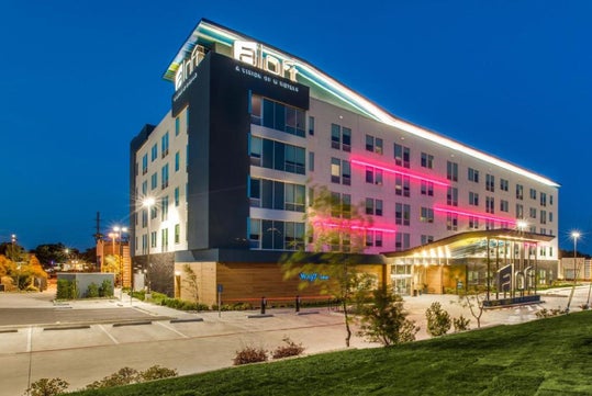 A modern, multi-story Aloft hotel building with illuminated signage and neon lights, viewed from the parking lot at dusk.