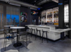 Modern bar with high tables and stools, abstract art on walls, and a well-stocked backlit bar area.