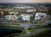 Aerial view of a suburban area with multiple large buildings, parking lots, and roads. The landscape includes green spaces and a body of water in the foreground.