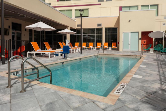 Outdoor swimming pool with orange lounge chairs and umbrellas on a tiled deck beside a beige building. Pool depth markers read "4 FT 1.2 M.