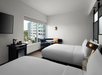 Hotel room with two double beds, wall-mounted TV, mini fridge, coffee maker, and a window view of modern buildings and a pool area.