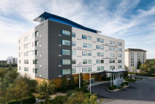 A modern six-story hotel building with a mix of white and gray exterior panels, surrounded by trees and a parking area under a partly cloudy blue sky.