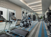 A modern gym with treadmills, elliptical machines, and weightlifting equipment. Walls have geometric patterns and mirrors, with overhead fluorescent lighting and a patterned carpet.