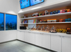 Self-serve snack station with shelves of chips, snacks, pastries, bananas, and a microwave. Large window and digital menu display above.