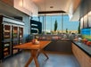 A modern hotel pantry with wooden table, beverage coolers, and shelves displaying snacks and drinks. Large windows offer a view of the parking lot and greenery outside.