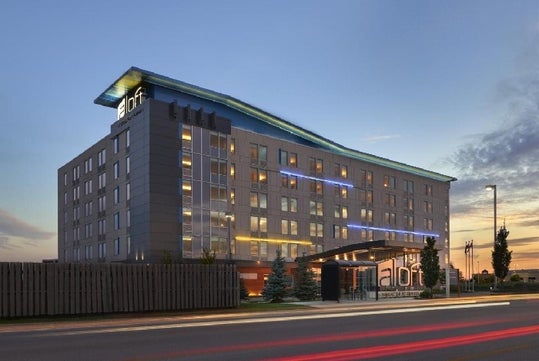 Modern hotel building at dusk with illuminated windows and signage, featuring a sloped roof and a driveway entrance in the foreground.