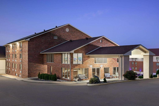 A brick hotel building with a covered entrance and large windows at dusk, surrounded by a paved driveway and landscaped shrubs.