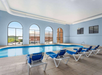 Indoor swimming pool area with several blue lounge chairs, arched windows with a view outside, and tiled flooring.