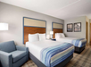 Hotel room with two double beds, a blue armchair, and a bedside lamp. Walls have framed photos, and bedding features blue-striped accents.