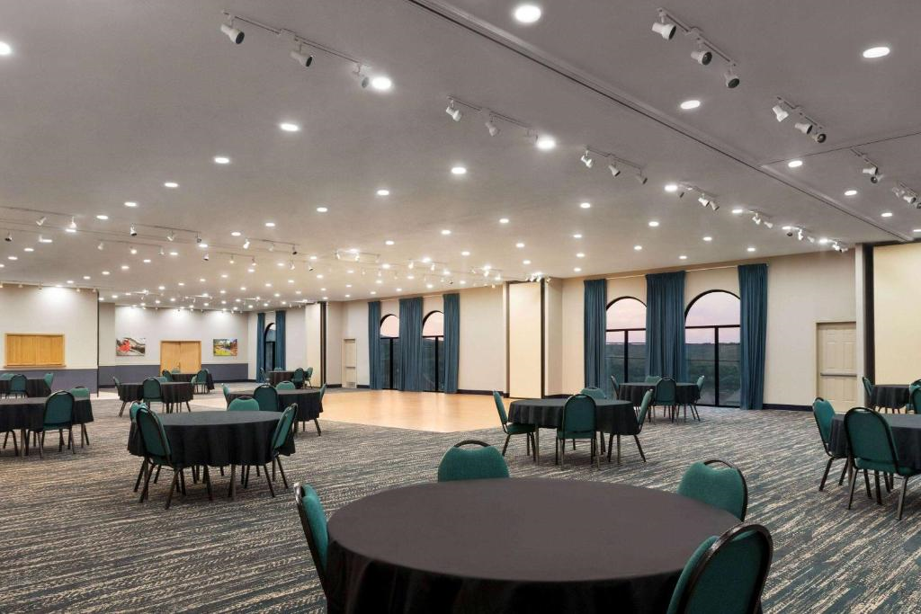 A large, well-lit conference room with round tables and green chairs, featuring high ceilings and windows with blue curtains.
