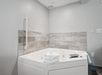 A white corner bathtub with jets is accompanied by a folded towel and toiletries against a gray tiled wall.
