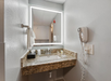 A hotel bathroom sink with a granite countertop, a backlit mirror, a mounted hairdryer, towel, and toiletries on the counter.