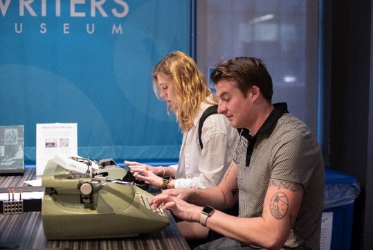 A man and woman sitting next to each other typing on old green typewriters  at the Story of the Day exhibit at the American Writers Museum.