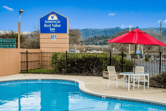 A brigth blue outdoor pool with a table with a red umbrella next to it and a sign for the Americas Best Value Inn in the backgorund.