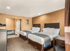 A hotel room with two queen beds, a table and chair, a dresser, and a bathroom on the back wall.