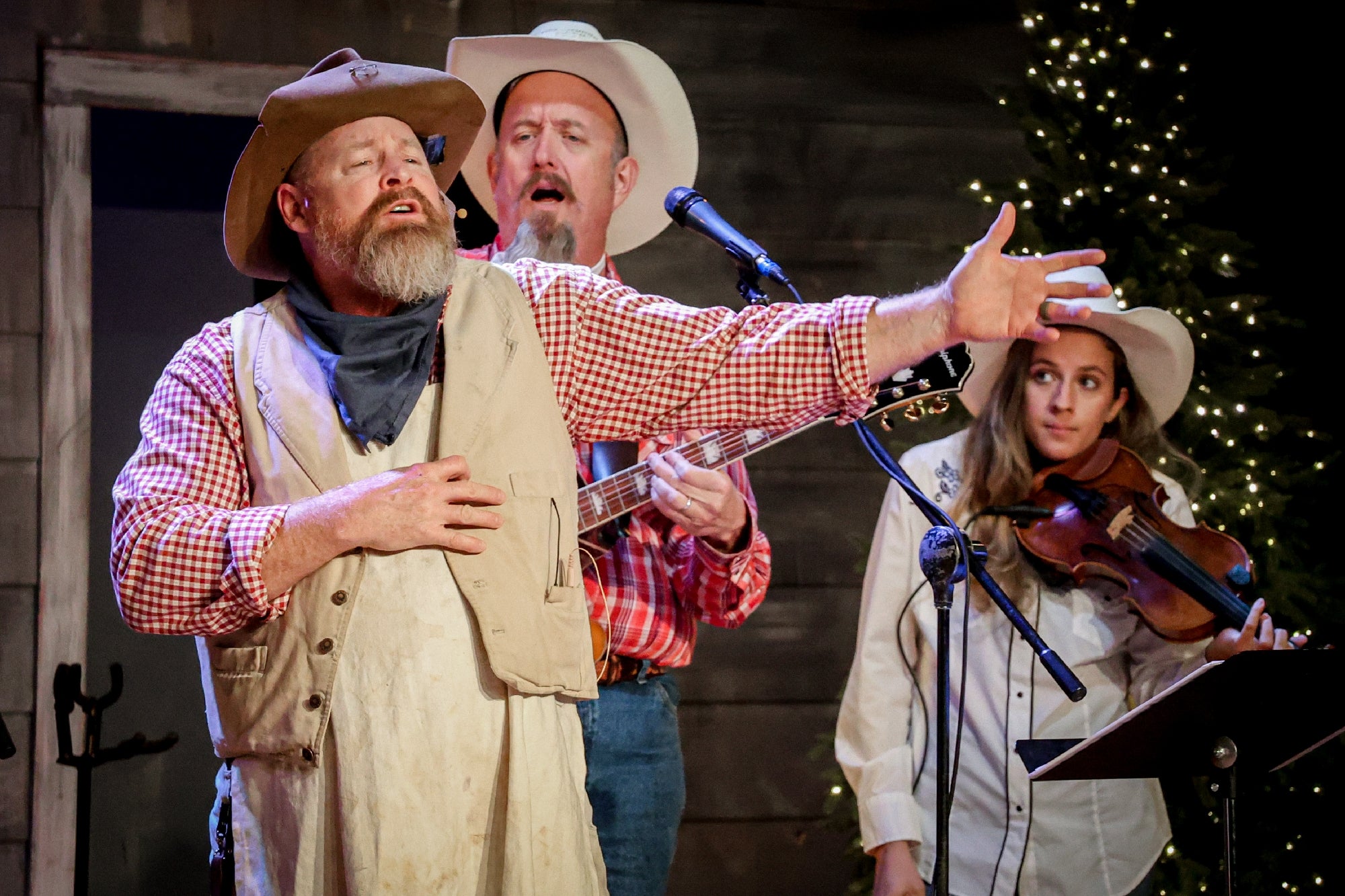 Three musicians in cowboy hats perform on stage; one sings expressively, another plays guitar, and the third plays violin. Christmas lights are visible in the background.