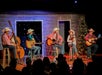 Five musicians wearing cowboy hats perform on stage with guitars, a double bass, and a fiddle, in front of a rustic backdrop and a decorated Christmas tree.