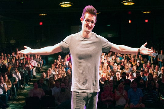 A man in a gray t-shirt stands on stage with arms outstretched, facing an audience seated in a darkened theater.