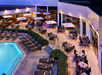 Outdoor hotel patio with tables, chairs, loungers, and umbrellas next to a lit pool area in the evening.
